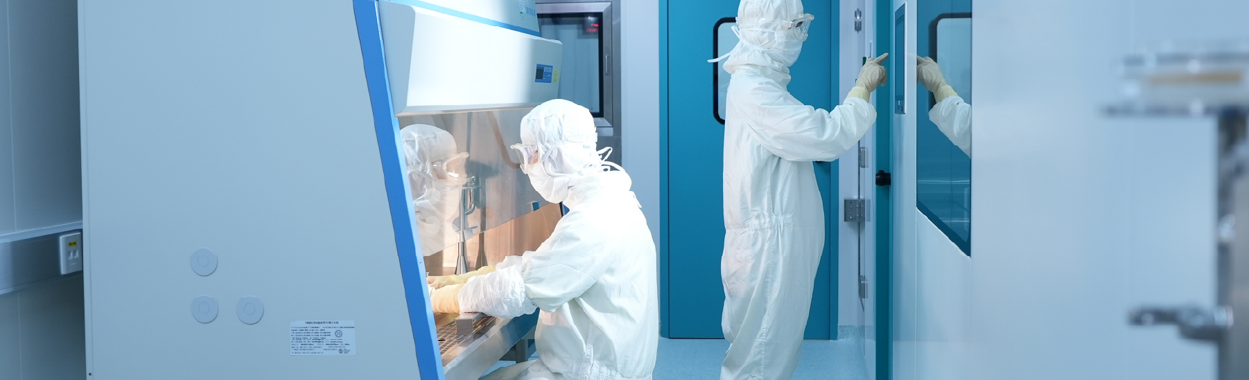 Scientists in full protective gear diligently service sophisticated machinery in a cleanroom; one operates equipment inside a fume hood, while another meticulously navigates a touchscreen, ensuring every step of the research pipeline is seamless and precise.