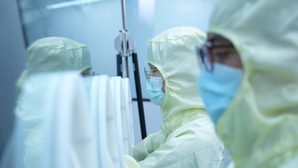 Two individuals in protective suits and masks meticulously work in a cleanroom environment, focusing on enhancing transduction efficiency behind glass panels to ensure optimal safety.